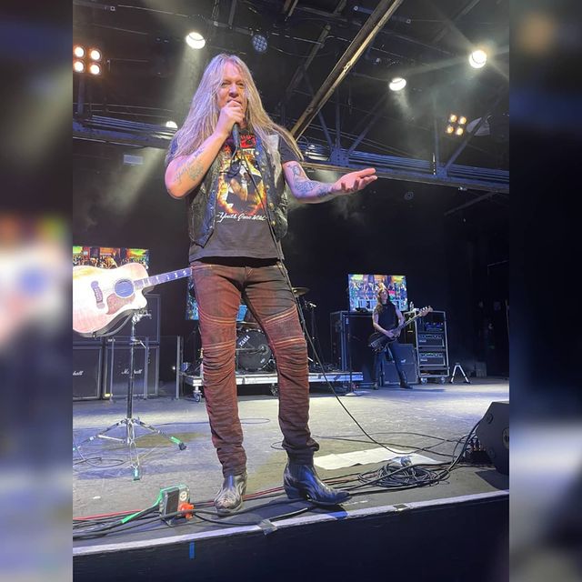 Sebastian Bach in a grey t-shirt and brown pants singing on the stage.